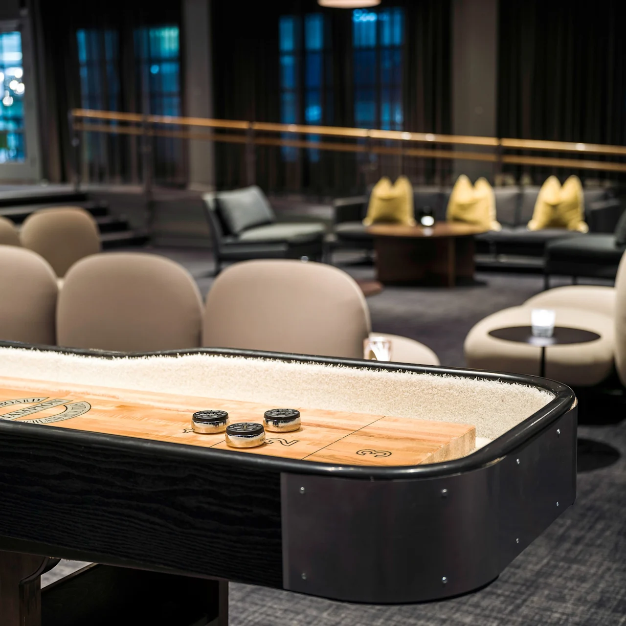 A shuffleboard table with pucks is in use in a modern lounge with plush seating and a carpeted floor. Text on the table reads: “SHUFFLEBOARD 9.”