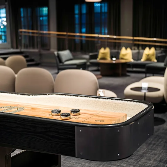 En shuffleboard-bord med brikker brukes i en moderne salong med komfortable sitteplasser og et teppebelagt gulv. Teksten på bordet lyder: "SHUFFLEBOARD 9."