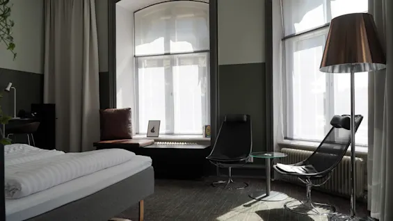 A modern hotel room features a neatly made bed, two sleek chairs, and a floor lamp. Sunlight streams through large windows, casting shadows on the carpeted floor.