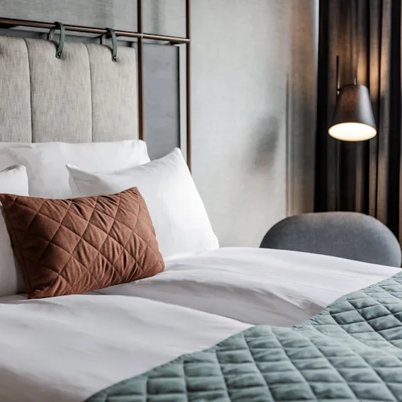 A comfortable hotel bed with white pillows, a brown accent pillow, and a teal blanket, ready for a relaxing stay.