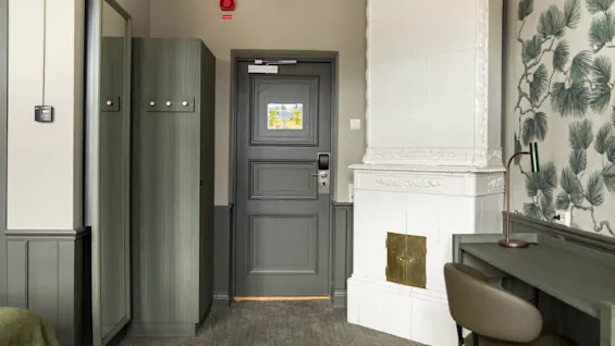 Kutsuva hotellihuoneen sisäänkäynti, jossa on vaatekaappi, klassinen kaakeliuuni ja toimiva työpöytäalue.