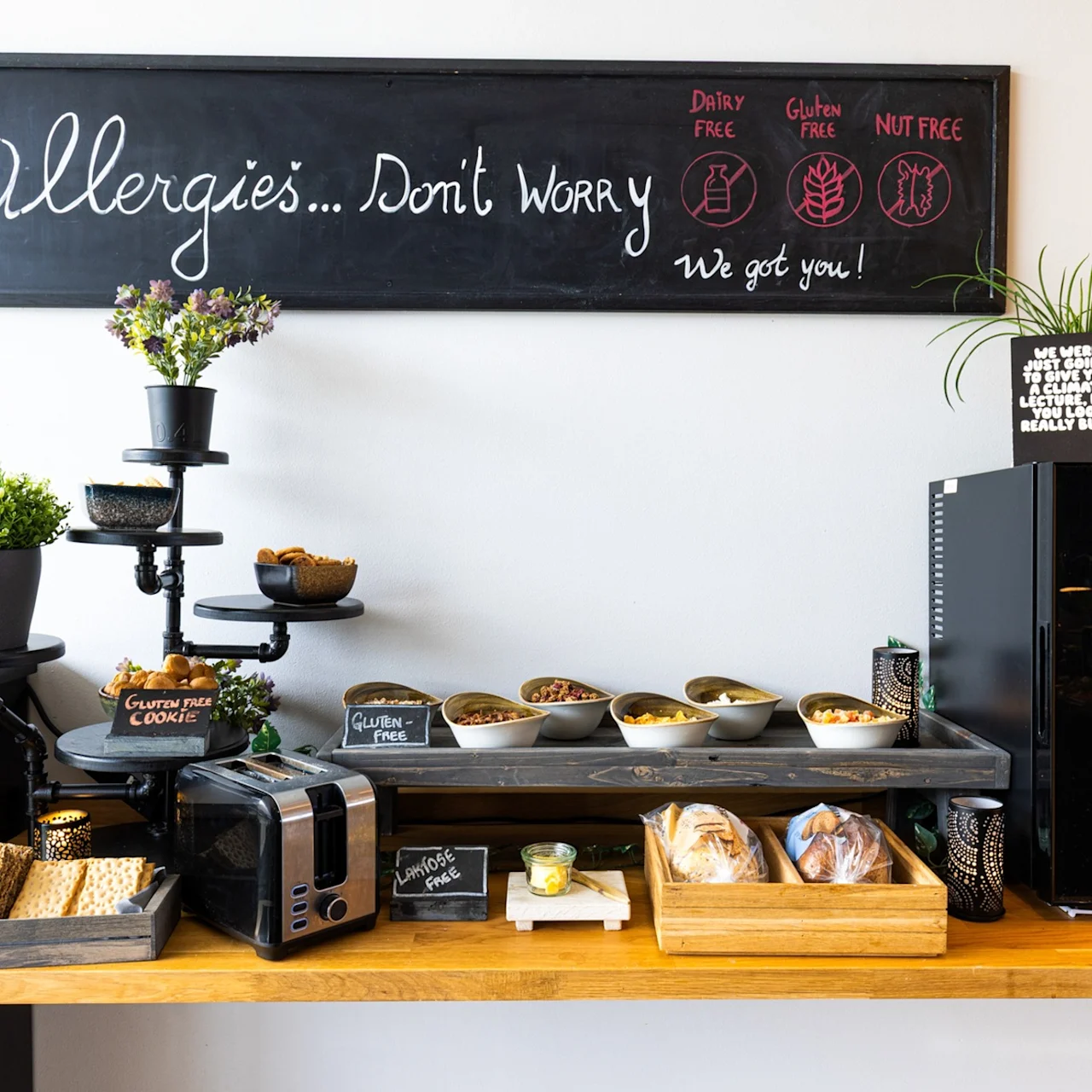 A breakfast buffet features cereals, a toaster, bread, and baked goods on a wooden counter. A blackboard above reads, "Allergies...Don't Worry. We got you! Dairy free, Gluten free, Nut free."