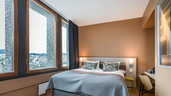 A neatly made double bed with gray linens sits in a warmly painted room, beside large windows overlooking a distant cityscape, enhancing the cozy, modern atmosphere.