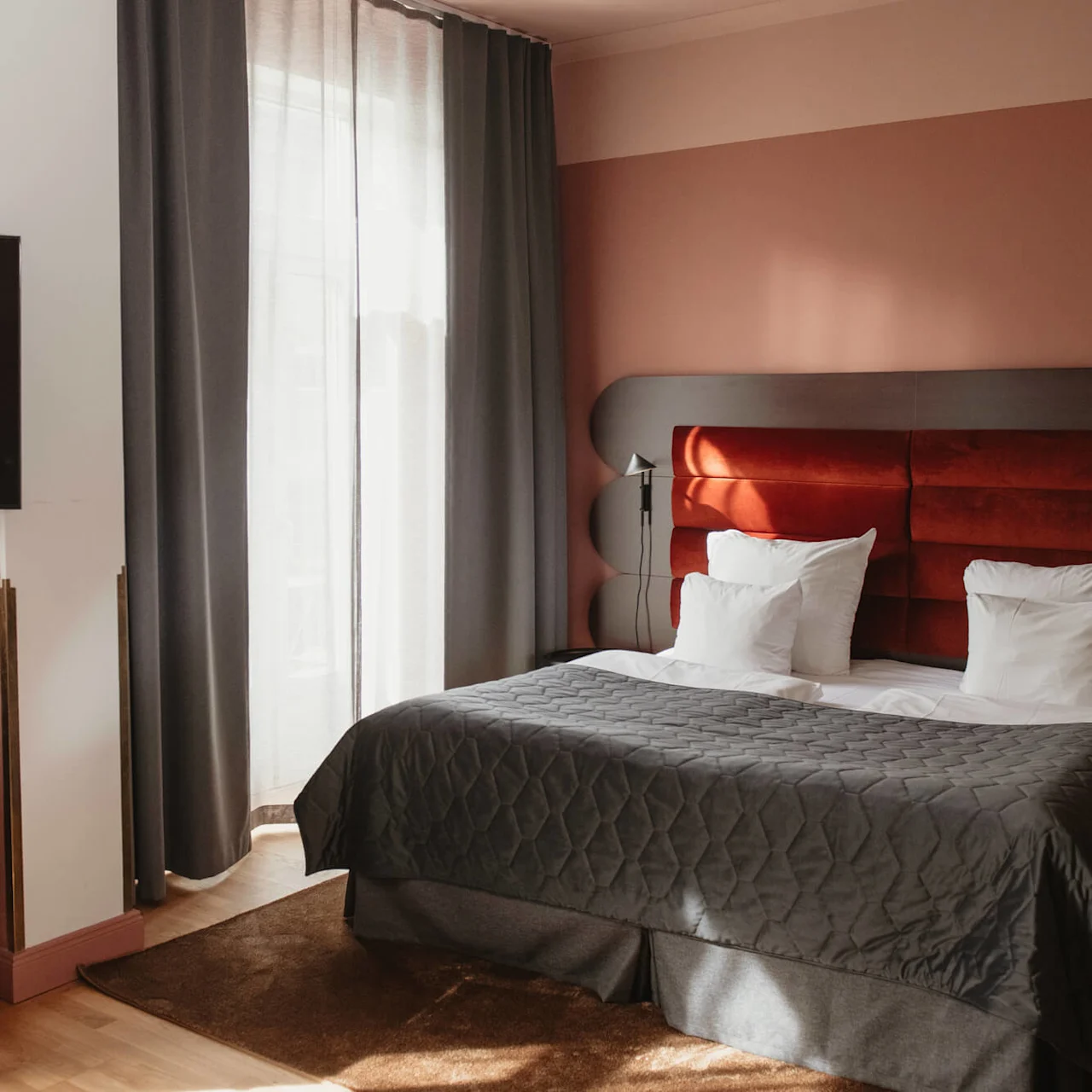 A neatly made bed with white and gray linens stands in a warmly lit room, with a dark curtain and a wall-mounted TV above a minibar. Text: "Retro."