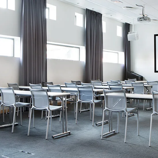 Grey chairs sit neatly arranged around tables in a bright, modern conference room. A large screen and projector are at the front, with natural light streaming through high windows.