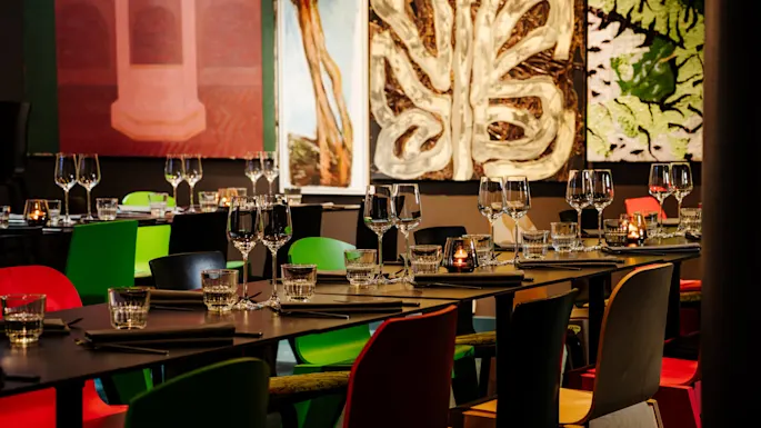 Bord dekket med vinglass og lys i en moderne restaurant, omgitt av fargerike stoler og abstrakt kunst på veggene.