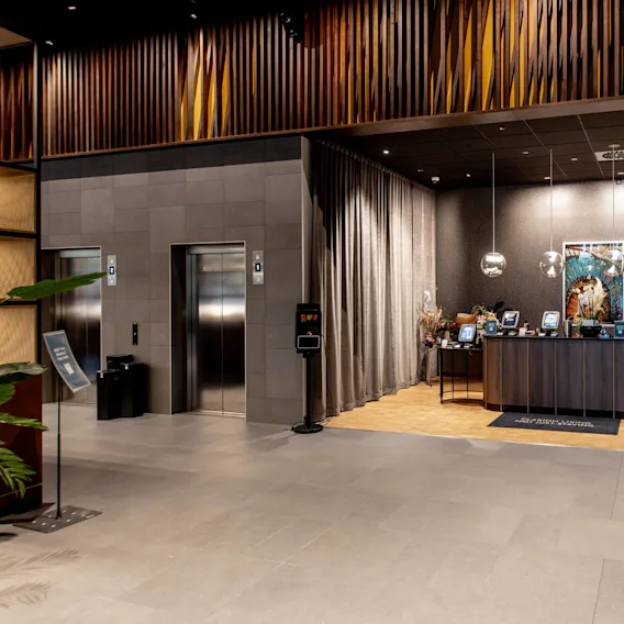 Reception desk with hanging lights, two computer monitors, and a decorative lion painting in a modern lobby. Adjacent, two elevators with metallic doors are framed by gray walls.