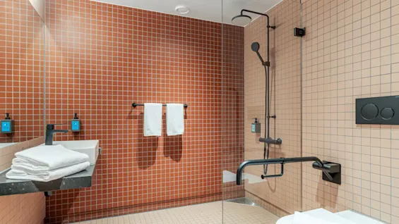 Et moderne badeværelse med håndvask og bruser, orange flisevægge, spejl, hvide håndklæder, toilet og minimalistiske sorte armaturer, inklusive håndtag og sæbedispensere.