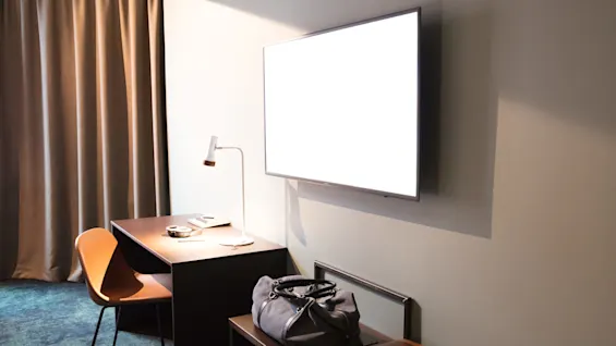 A wall-mounted TV emits light in a hotel room. A desk with a lamp, chair, and papers is nearby. A duffel bag rests on a bench under the TV.