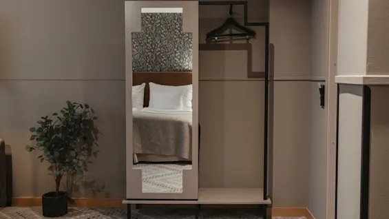 En modern hotellrumsinredning med en spegel som reflekterar en prydligt bäddad säng, en klädställning och en krukväxt.