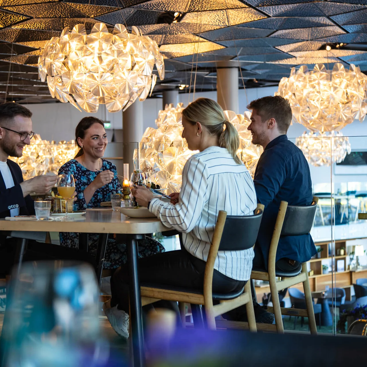 Fire personer sidder ved et spisebord, nyder et måltid og samtale i en moderne restaurant, oplyst af store, dekorative pandelamper.