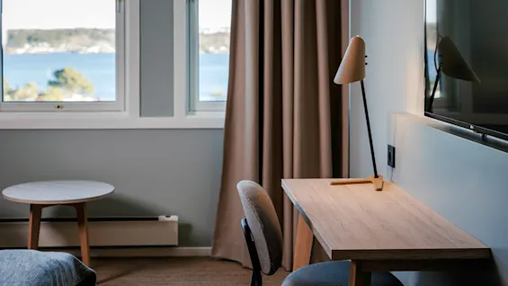 A wooden desk with a modern lamp sits beside a window showing a scenic ocean view. Nearby, a small round table and a chair complete the room's cozy interior.