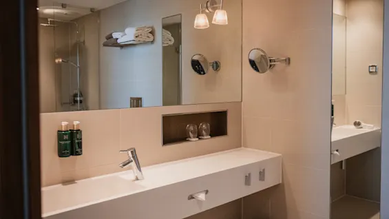 Sink with faucet and toiletries, reflected in a large mirror, in a beige-colored modern bathroom. Towels are neatly stacked on a shelf, and shower visible in the background.