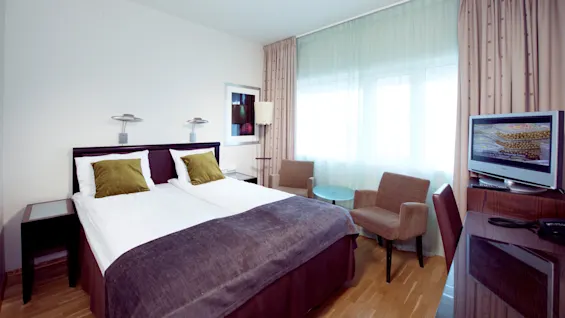 A tidy hotel room with two beds, side tables, a TV, armchair, and table by a brightly lit window with sheer curtains.