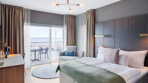 Et pent oppstilt hotellrom har en stor seng med puter og teppe, et skrivebord med glass, og en grå sofa. Balkongdørene åpner mot en vannutsikt med stoler utenfor.