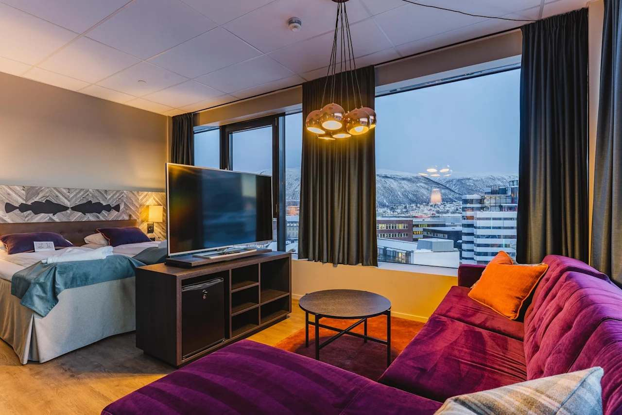 Viihtyisä hotellihuone, jossa on iso sänky, televisio ja violetti sohva, tarjoten luonnonkauniin näkymän lumisille vuorille ja kaupunkiin.