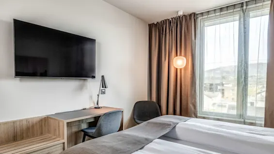 En pænt redt seng vender mod et vægmonteret TV i et moderne hotelværelse. Et skrivebord med en stol er i nærheden, og et vindue med gardiner giver naturligt lys.