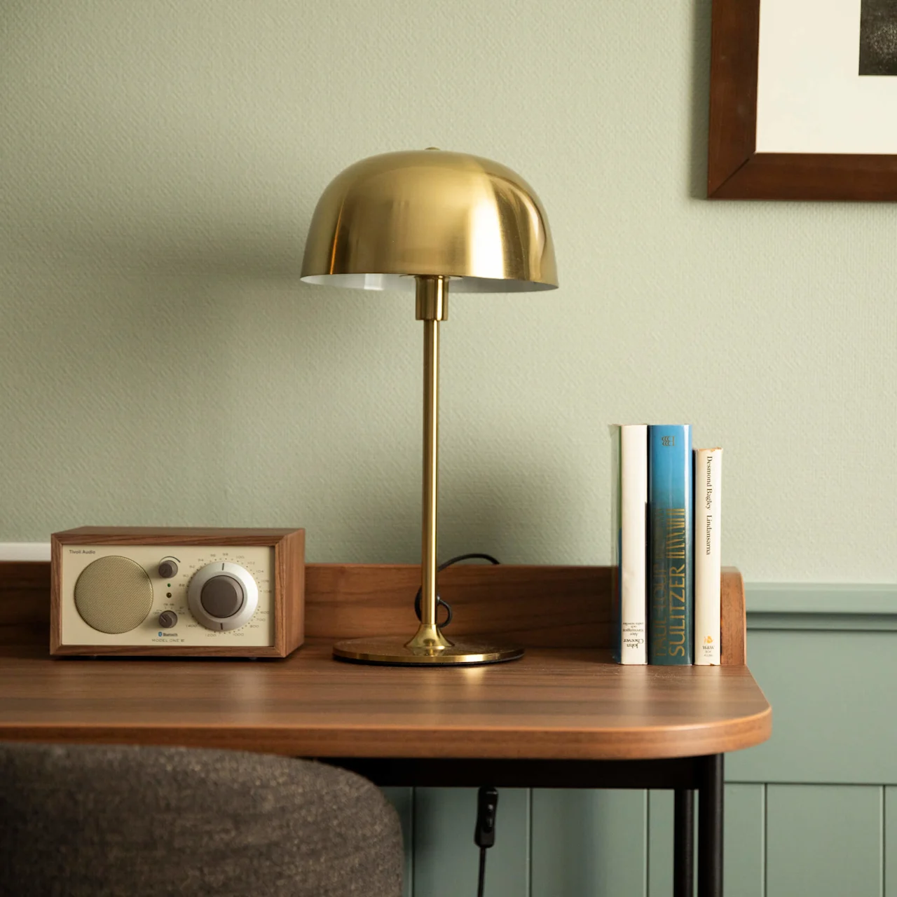 En mässingbordslampa står på ett träbord bredvid en vintage radio och tre böcker. En inramad bild hänger på den blekgröna väggen.