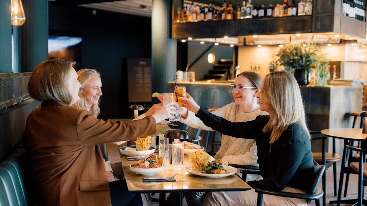 Four people sit at a restaurant table, clinking glasses in a celebratory toast. Plates of food and glasses surround them in a warmly lit dining area.
