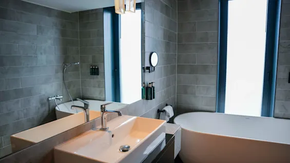 Modern bathroom featuring a wide white sink with a chrome faucet and a freestanding bathtub. Gray tiled walls surround the space, with a large mirror and a frosted window enhancing brightness.