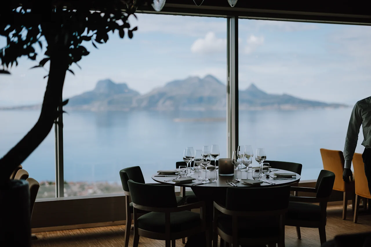 Et bord med flot arrangeret glas og bestik har panoramaudsigt over bjerge og vand gennem et stort vindue. En person står i nærheden i en restaurantsammenhæng.
