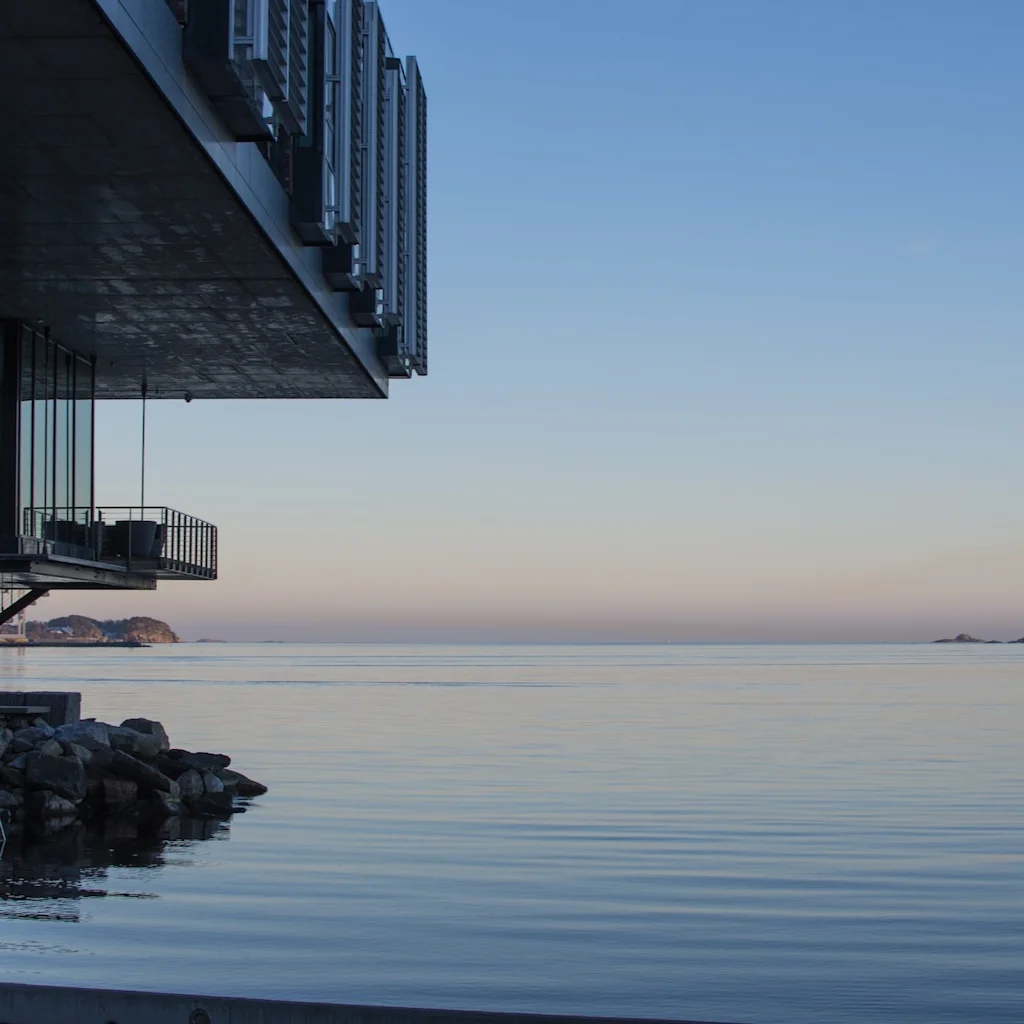 En modern byggnad med en balkong som sträcker sig över lugnt vatten, mot en fridfull himmel.