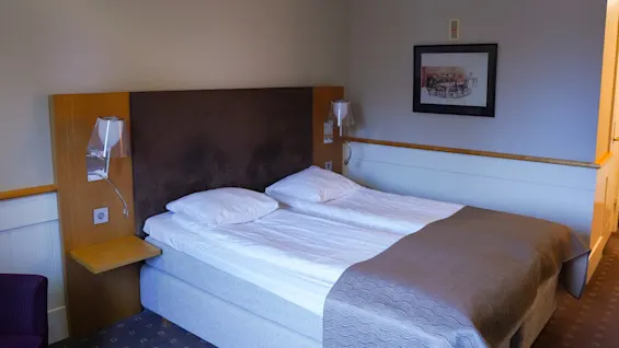 A neatly made twin bed in a hotel room with a wooden headboard, a nightstand, a wall lamp, and artwork.