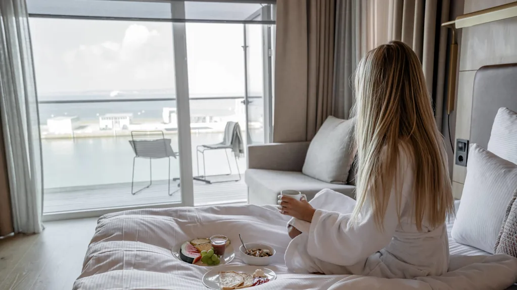 A woman in a white robe sits on a bed, holding a mug, with breakfast plates nearby. She gazes out a window overlooking water and buildings.