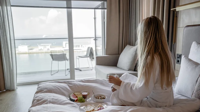 A woman in a white robe sits on a bed, holding a mug, with breakfast plates nearby. She gazes out a window overlooking water and buildings.