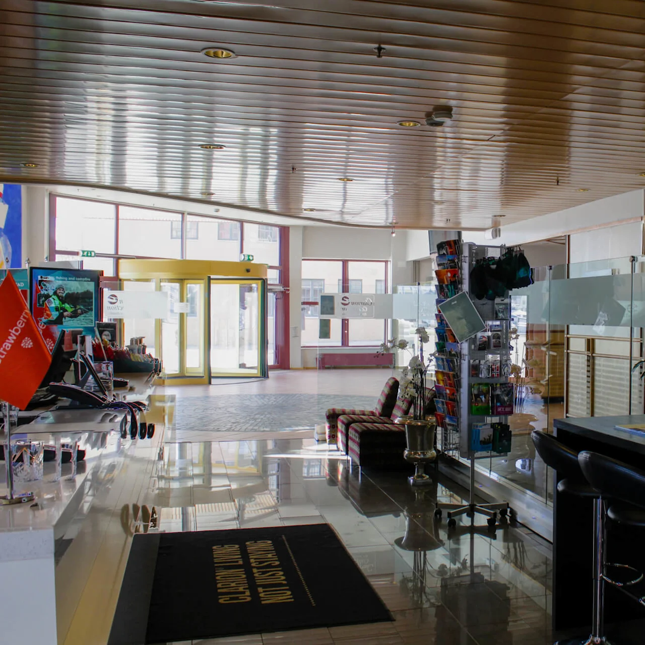 En moderne lobby med glasdøre, højt loft og en reception med brochurer og lys, omgivet af siddepladser og dekorationer. Tekst på måtten lyder "CHILDREN PLAYING NO LUGGAGE STORAGE".