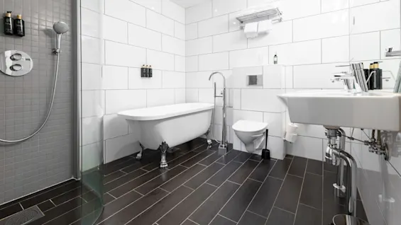 Bathtub stands in modern bathroom, surrounded by white tiles and dark wood flooring. Nearby, a toilet, sink, shower, and towel rack complete the sleek, minimalist design.