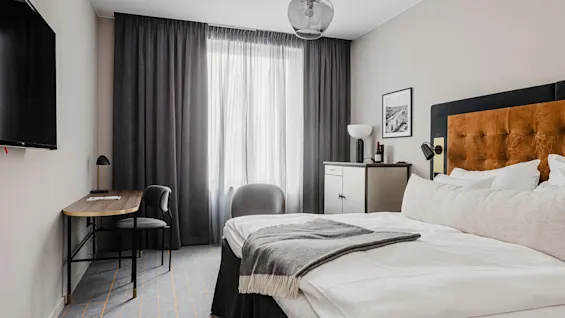 En pænt redt seng med puder og tæppe i forgrunden, ved siden af et moderne skrivebord og stol, vendt mod et vindue med mørke gardiner i et minimalistisk rum.
