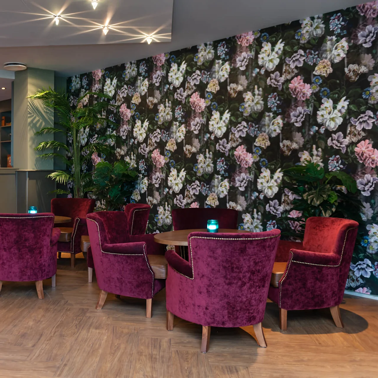 Plyschiga sammetstolar omger träbord, uppställda mot en blommig tapet i en mjukt belyst lounge, vilket skapar en elegant och mysig miljö.
