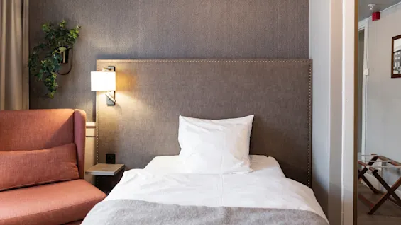 A welcoming hotel room at Home Hotel Grand Bodø featuring a neatly made bed, a plush armchair, and a decorative plant.