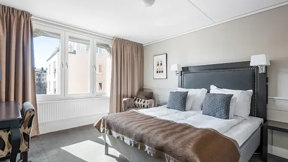 A neatly made bed with pillows against an upholstered headboard, flanked by lamps, in a bright, modern bedroom with large windows and curtains.