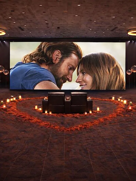 En romantisk filmuppsättning med en stor skärm som visar ett par, två sittplatser och ljus som bildar ett hjärta på golvet.