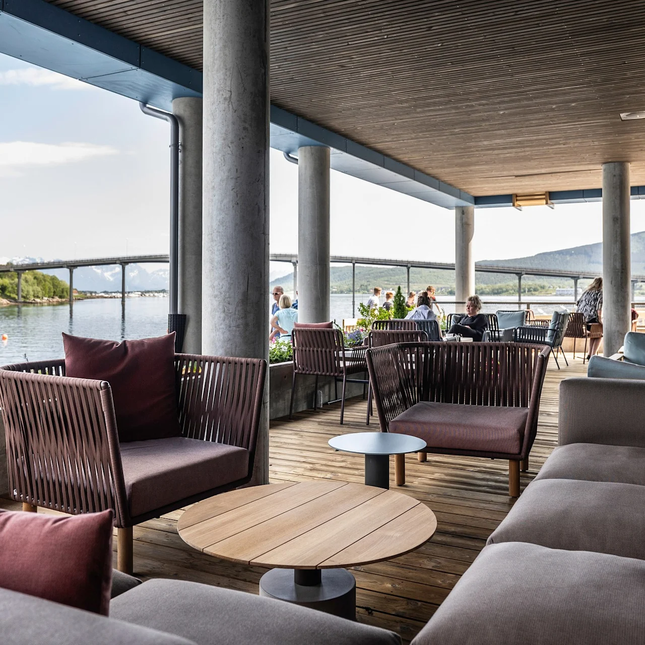 Komfortable udemøbler med borde arrangeret under en overdækket terrasse med udsigt over en naturskøn bugt og fjerne bjerge, der giver et afslappende sted for besøgende at nyde udsigten.