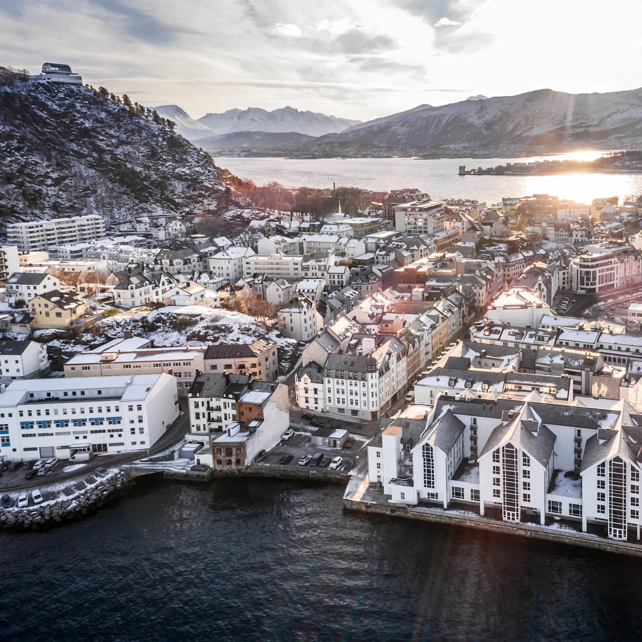 En kystby med snedækkede bygninger ligger ved en stor vandmasse. Omgivende bjerge rejser sig i baggrunden under en lys himmel.