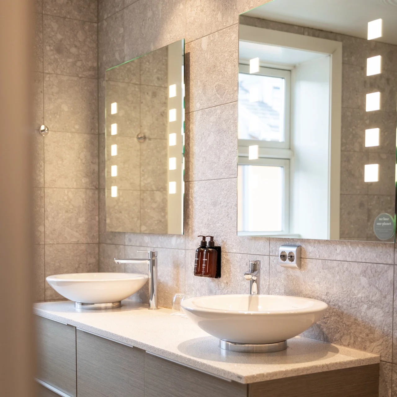 Två moderna vita handfat med kromkranar står på en beige bänkskiva under upplysta speglar. Grå kaklade väggar och ett fönster skapar en ljus, elegant badrumsmiljö.