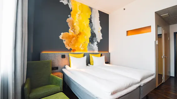 A double bed with white linens and yellow pillows is beside a green armchair. A wall mural of yellow, black, and white abstract design decorates the modern hotel room.