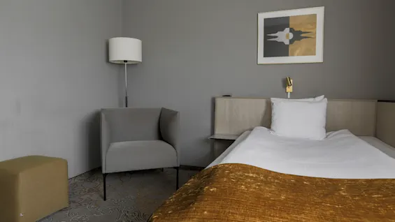 Bed with white pillows and golden blanket; beside it, a gray armchair and floor lamp in a minimalistic room with gray walls. A modern artwork hangs above.