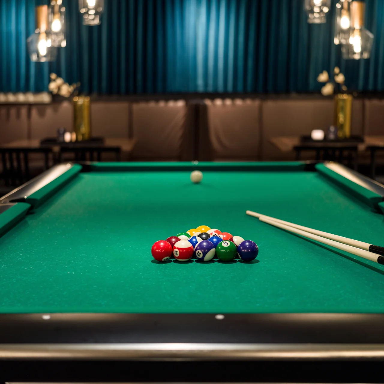 A pool table with balls arranged in a triangle is ready for play, set in a dimly lit room with cushioned seating and hanging lights in the background.