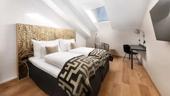 A neatly arranged attic bedroom featuring a double bed with patterned throw, a desk area with chair, under a slanted ceiling with a skylight.