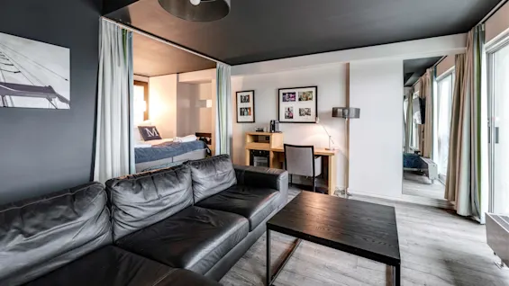 En svart lädersoffa vetter mot ett soffbord i ett modernt vardagsrum med grå väggar. En säng syns delvis bakom gardiner; ett skrivbord och foton pryder väggen.