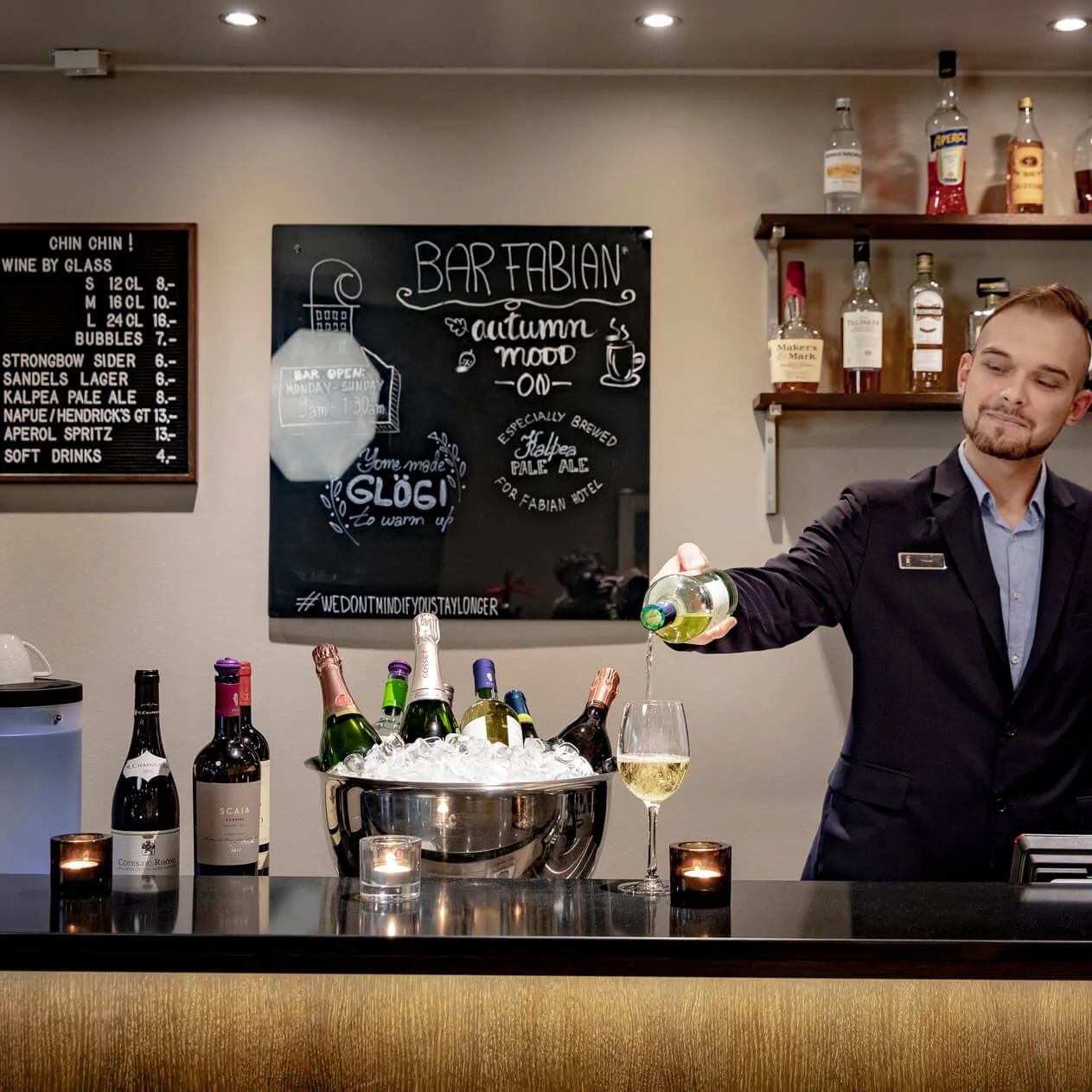 En bartender heller vin i et glass på en bar med hyller av brennevinsflasker. Tavla sier: "BAR FABIAN, autumn mood on, specially brewed Fabian pale ale" og mer. Vinliste: "CHIN CHIN!"