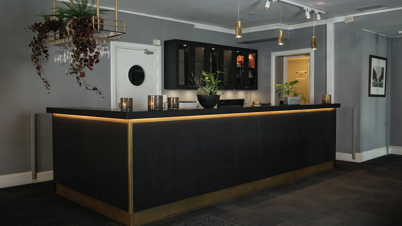 A modern reception desk with wooden accents stands in a dimly lit room adorned with plants on the counter. Wall-mounted cabinets and decorative lighting complete the sleek interior design.