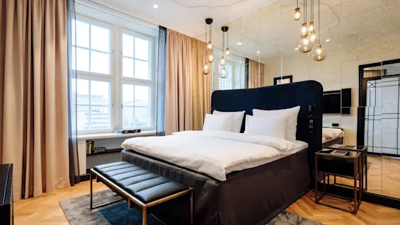 A neatly made bed with white linens stands against mirrored wall panels, lit by hanging pendant lights. Surrounding are elegant curtains, a window, and a wooden herringbone floor.