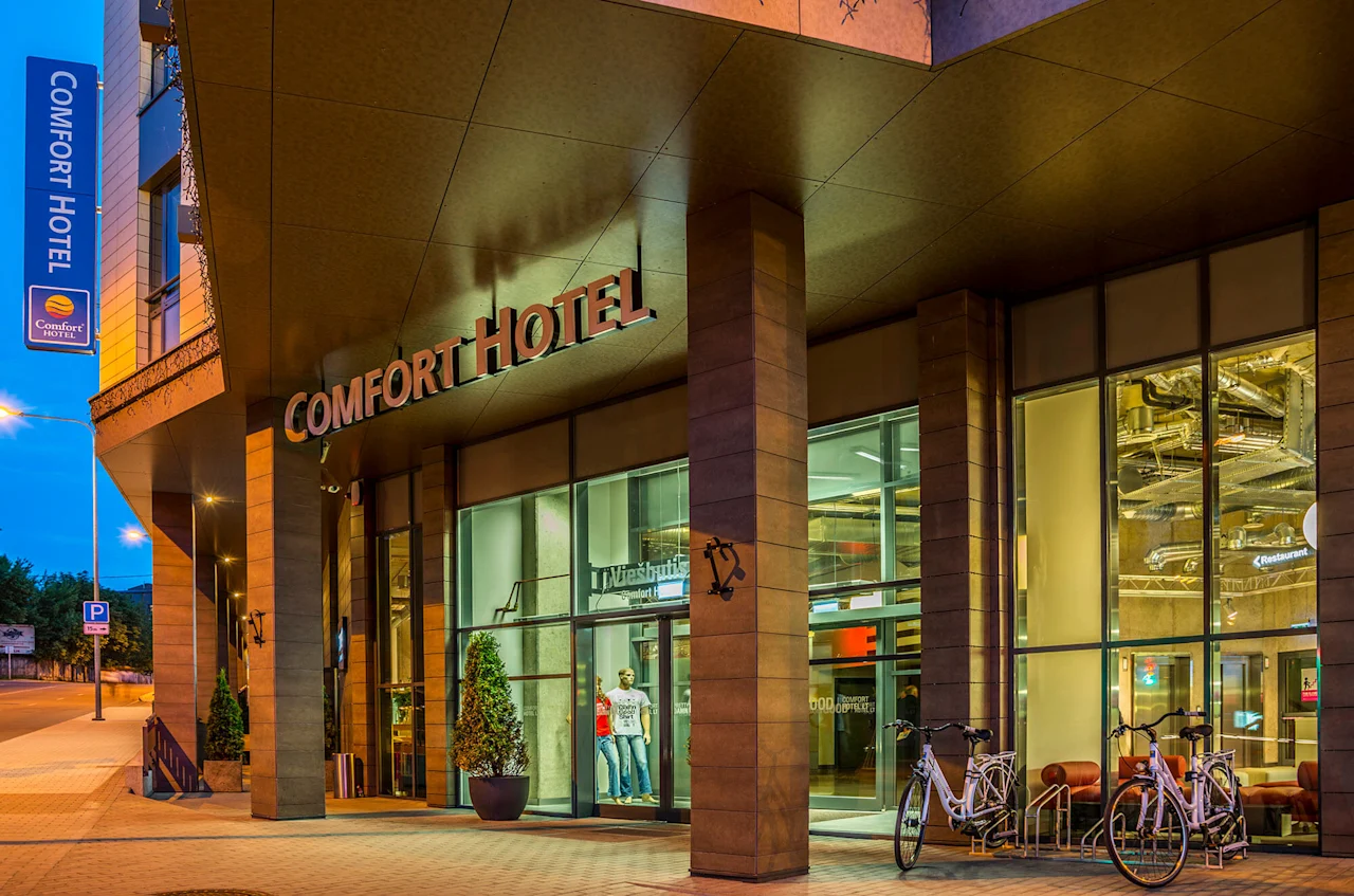 The entrance of a Comfort Hotel, with bicycles parked outside and a modern glass facade. Signs read "Comfort Hotel" and "Restaurant." Exterior is lit at dusk.  