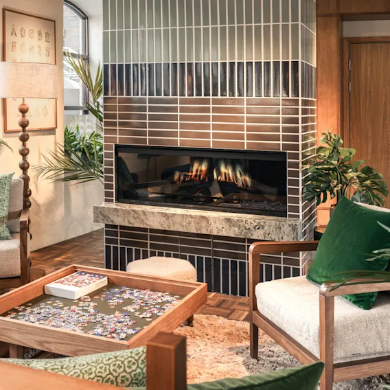 A warm and inviting living space featuring a lit fireplace, comfortable armchairs, and a jigsaw puzzle on a coffee table. ABCDE FGHIJ.
