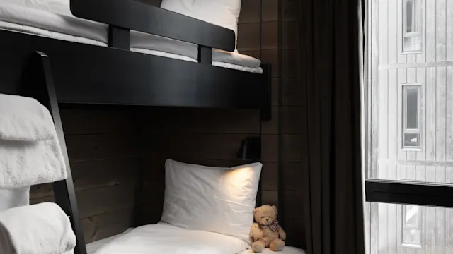 Bunk beds in a wooden-paneled room with a teddy bear on a lower bed. White towels hang on a black ladder, near a large window covered by a dark curtain.